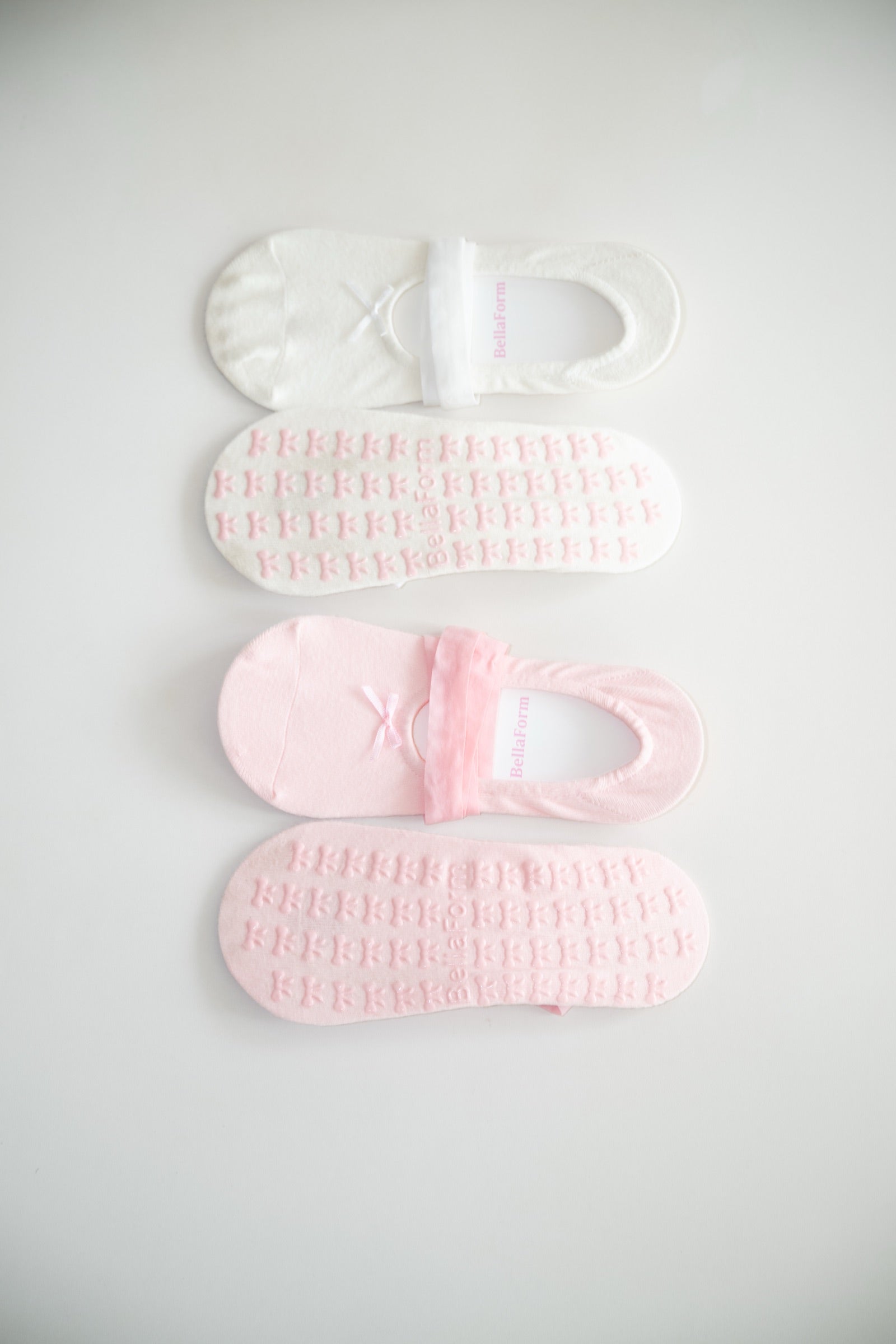BellaForm grip socks in both colorways side by side, Ballet Pink and Cloud White, showing the knit top and the bow-print silicone sole of each.
